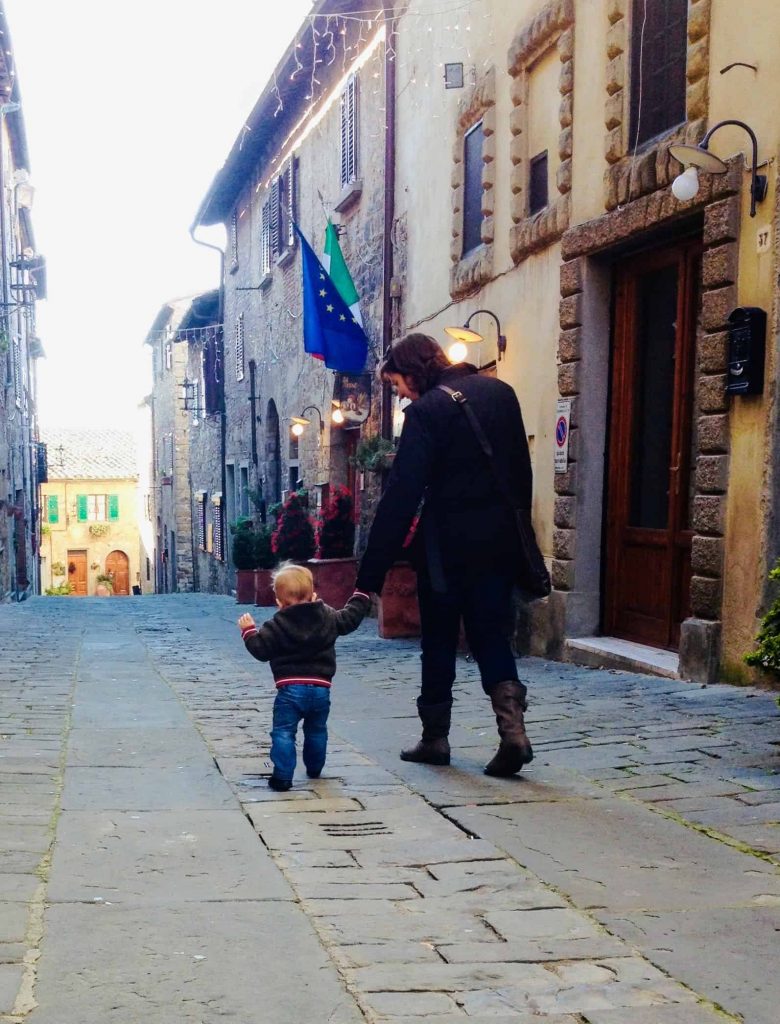 Woman and young son walking hand-in-hand away from the camera in a village in Tuscany, Italy.  There is an EU flag and an Italian flag hanging from the side of the building on the left.  The street is made of large stones. 