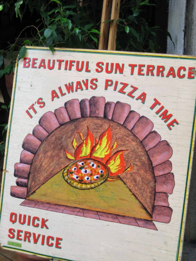 Painted wooden sign in front of bush (presumably outside a restaurant door) with 'beautiful sun terrace - it's always pizza time - quick service' painted on it.  In the middle of the sign is a painting of pizza being cooked in a wood fire oven.