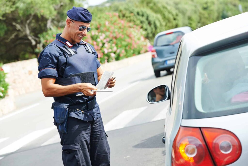 Italian carabinieri officer in uniform, which includes the berretto (hat), is smiling and checking the documents of a driver in a silver car.  