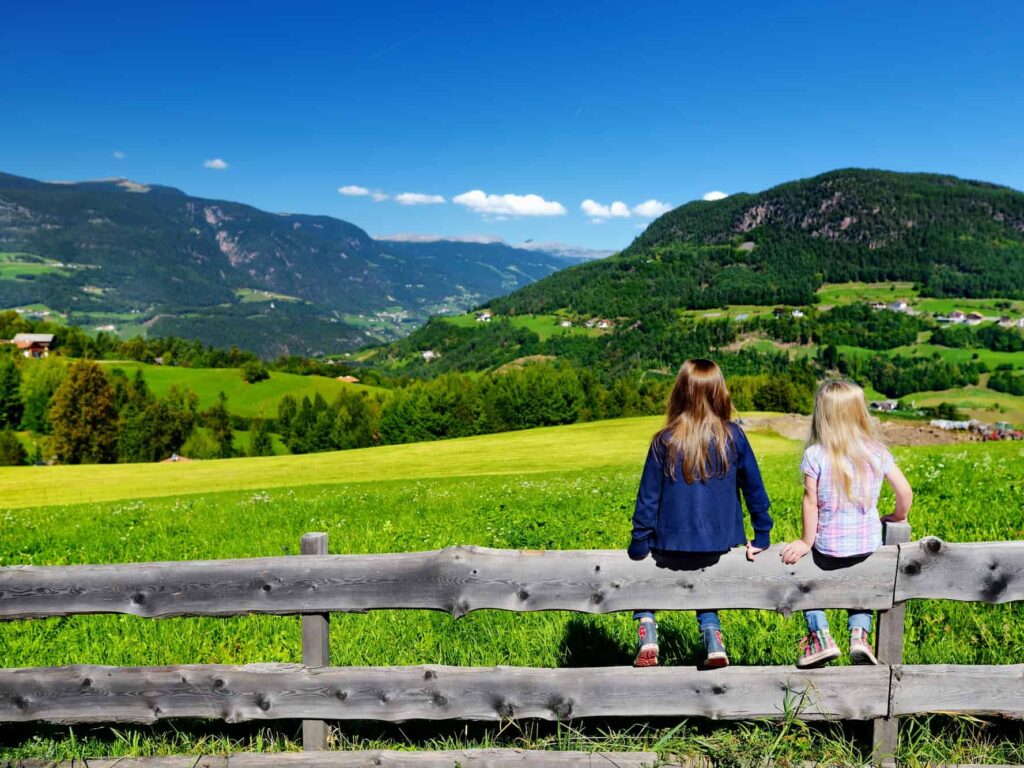 Sisters sitting on a wooden fence and looking out at the green hills and mountains in the distance.