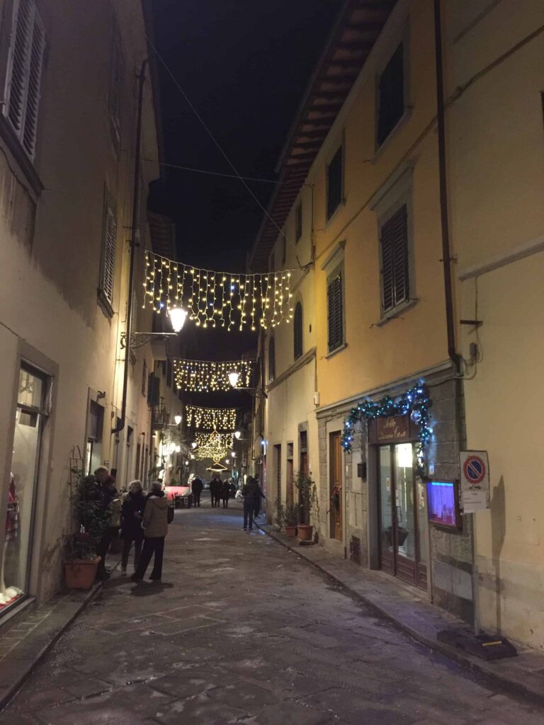 Lights decorate a street in a small village in Italy.  You can see a few people in coats.
