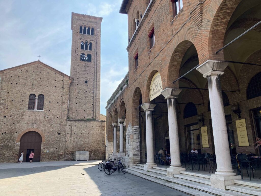 Outdoor seating under a portico and next to Basilica di San Francesco. Two people stand at door to basilica.