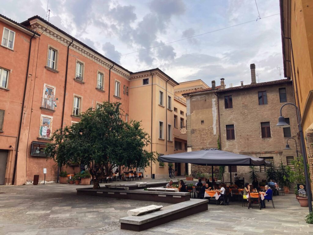 Outdoor seating at Osteria dei Battibecchi. Piazza surrounded by orange and brown buildings and people eating under a large brown umbrella. There is a large tree next to the restaurant in the piazza.