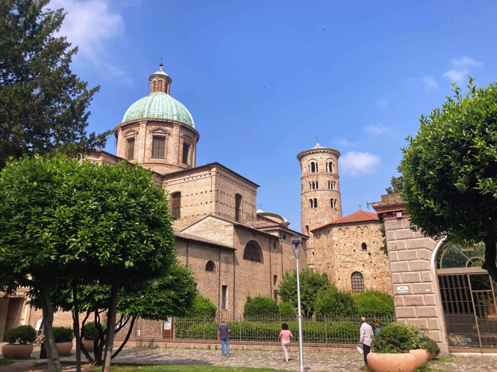 View of outside of Neonian Baptistry in Ravenna, Italy. There are green trees and bushes on the property and few people are walking on the cobblestone path in front.