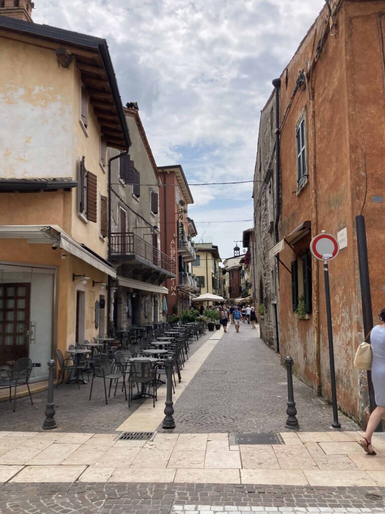 Narrow street in Lazise in Lake Garda on a sunny day. You can see tables and chairs on the left and people walking on the street in the distance.