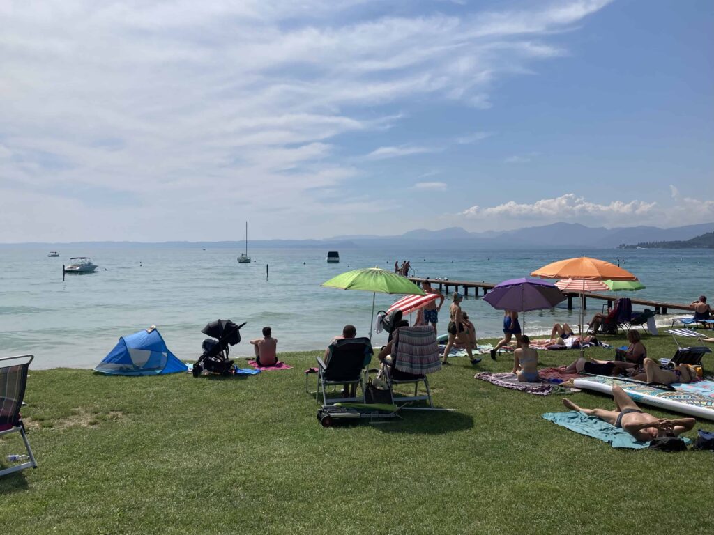 People sitting on a grassy shore on Lake Garda. Umbrellas and beach towels on grass. A few boats in the water.