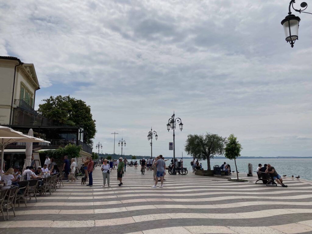 Striped lungolago in Lazise, Lake Garda. People walking on it, and some are sitting on benches on right and at outdoor cafes on left.