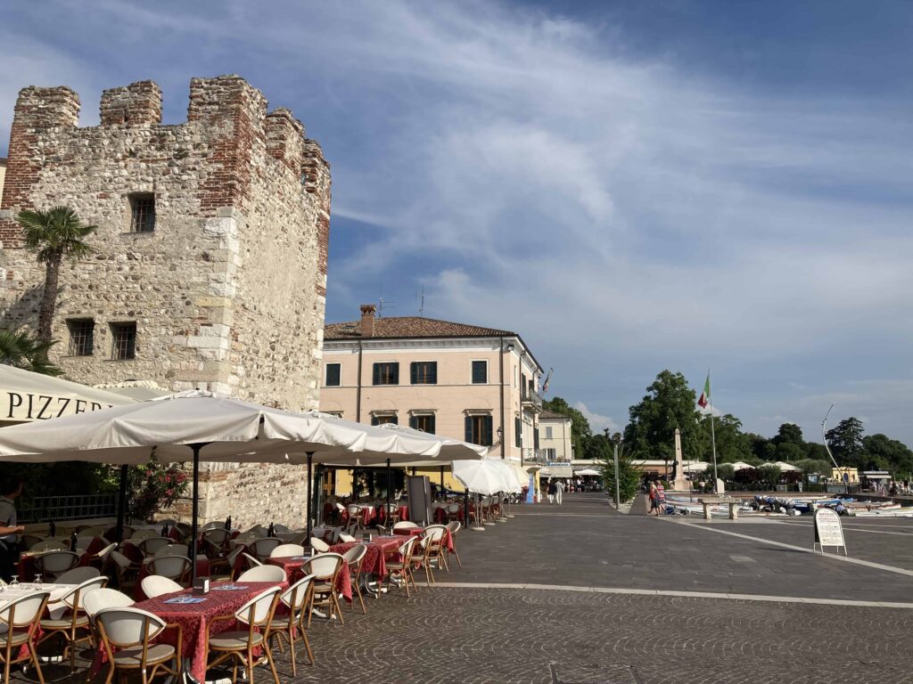 Tower on side of lakefront in Bardolino, Italy. Outdoor tables at restaurant on left.