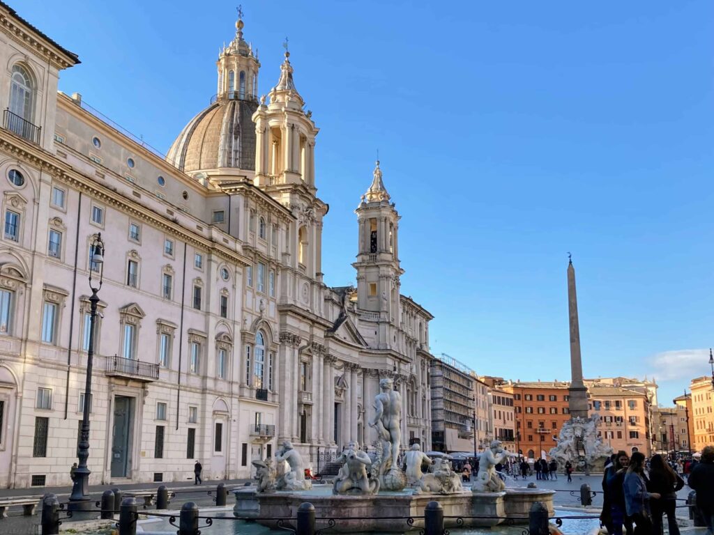 View of Piazza Navona in Rome on a quiet winter morning. There are a few people in the piazza in coats. You can see the marble fountains and statues and the buildings surrounding the square.
