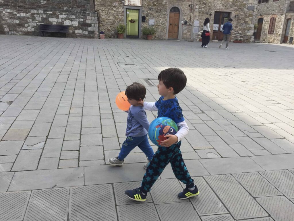 Two brothers walking together in an empty piazza in Italy. They're each carrying a ball.