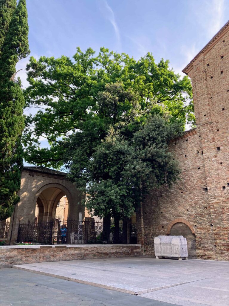 Dante's tomb by the Basilica of San Francesco in Ravenna. Leafy green trees overhead.