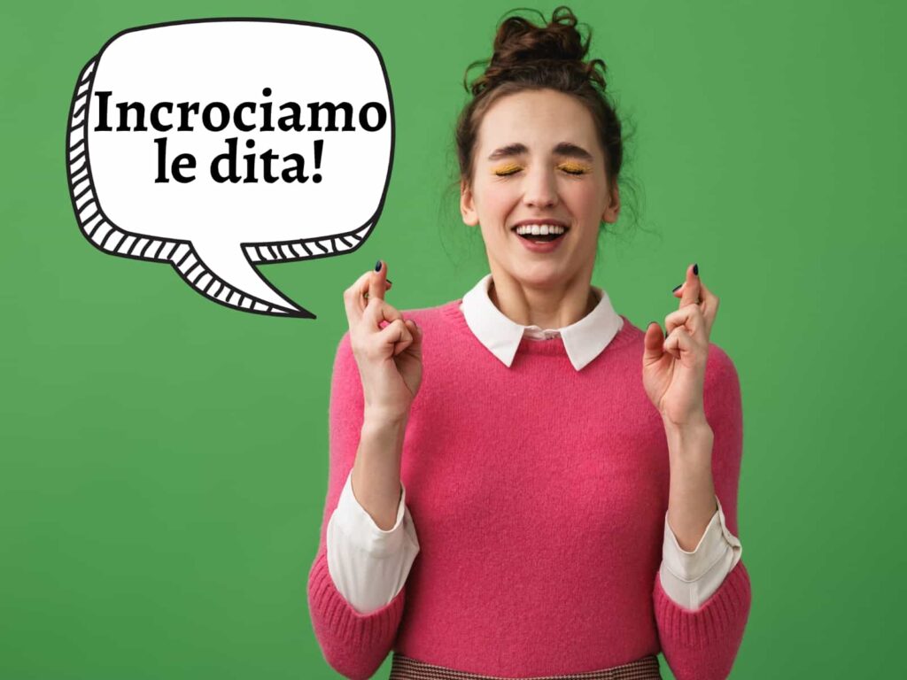 Woman in pink sweater closes eyes and crosses fingers and says in graphic speech bubble, 'Incrociamo le dita!'