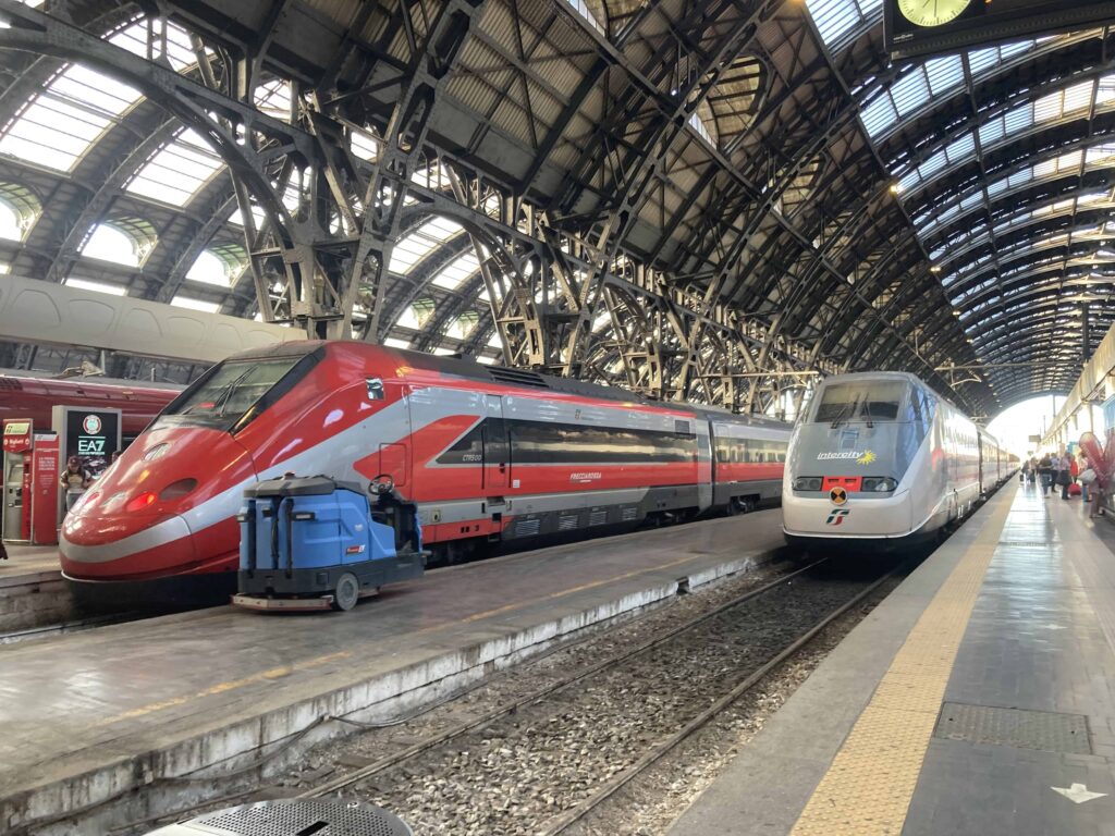 Two trains at the tracks in the Milano Centrale train station.