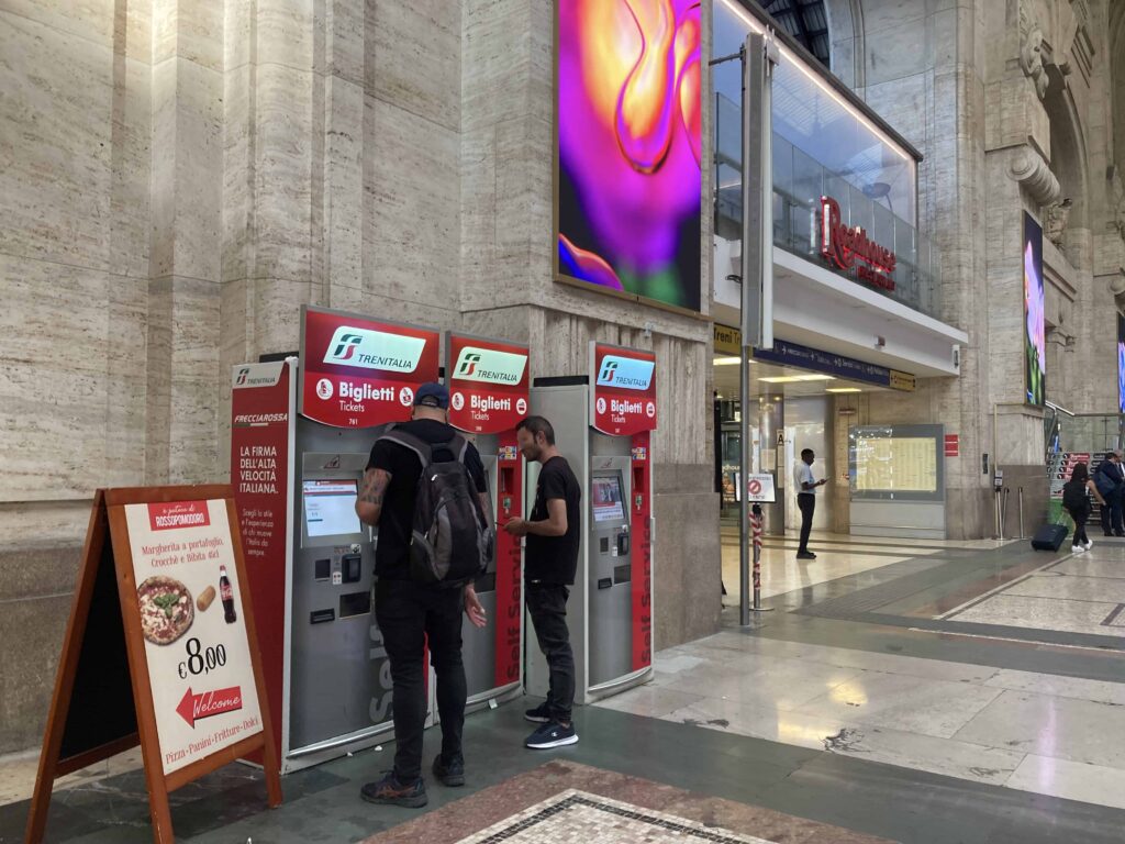 Two men buying tickets in red automatic ticket machines at Milan Central train station.