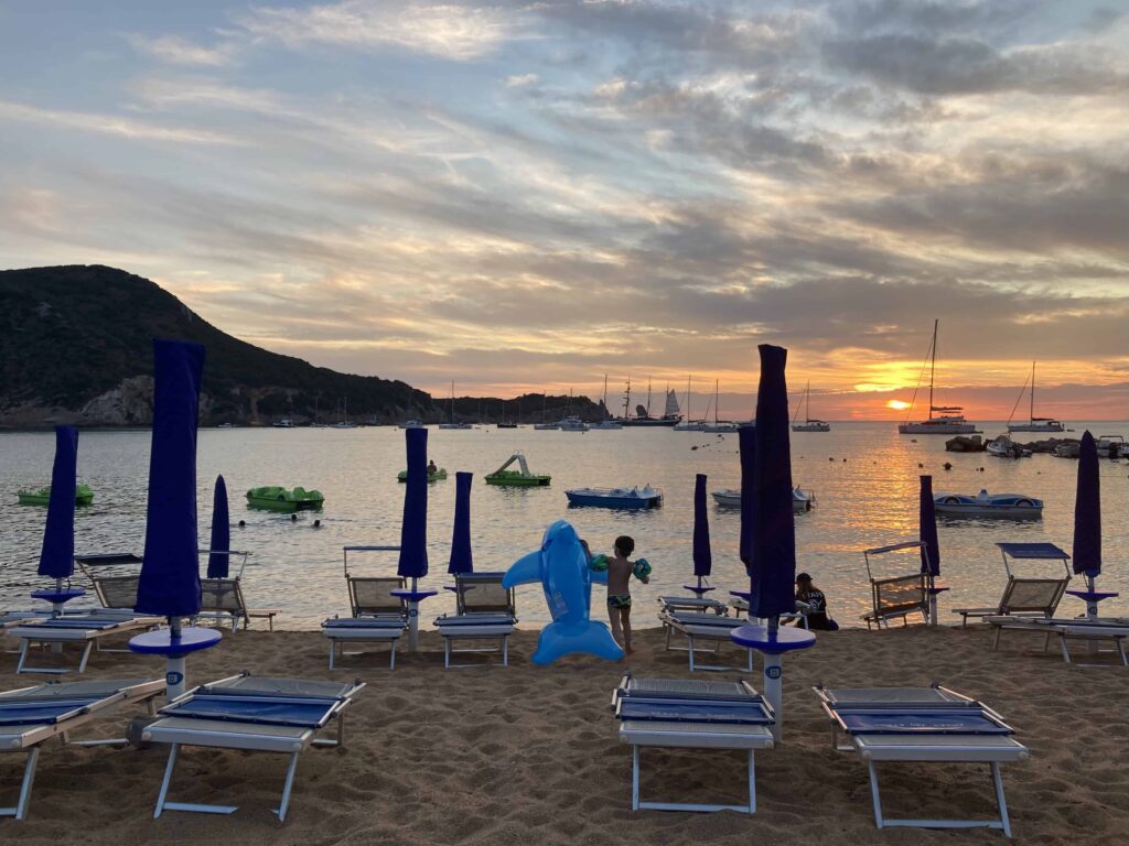 Sunset at private beach in Italy. You can see pedal boats in water, sailboats in distance. On beach, boy carries an inflatable dolphin past closed beach umbrellas and sun loungers.