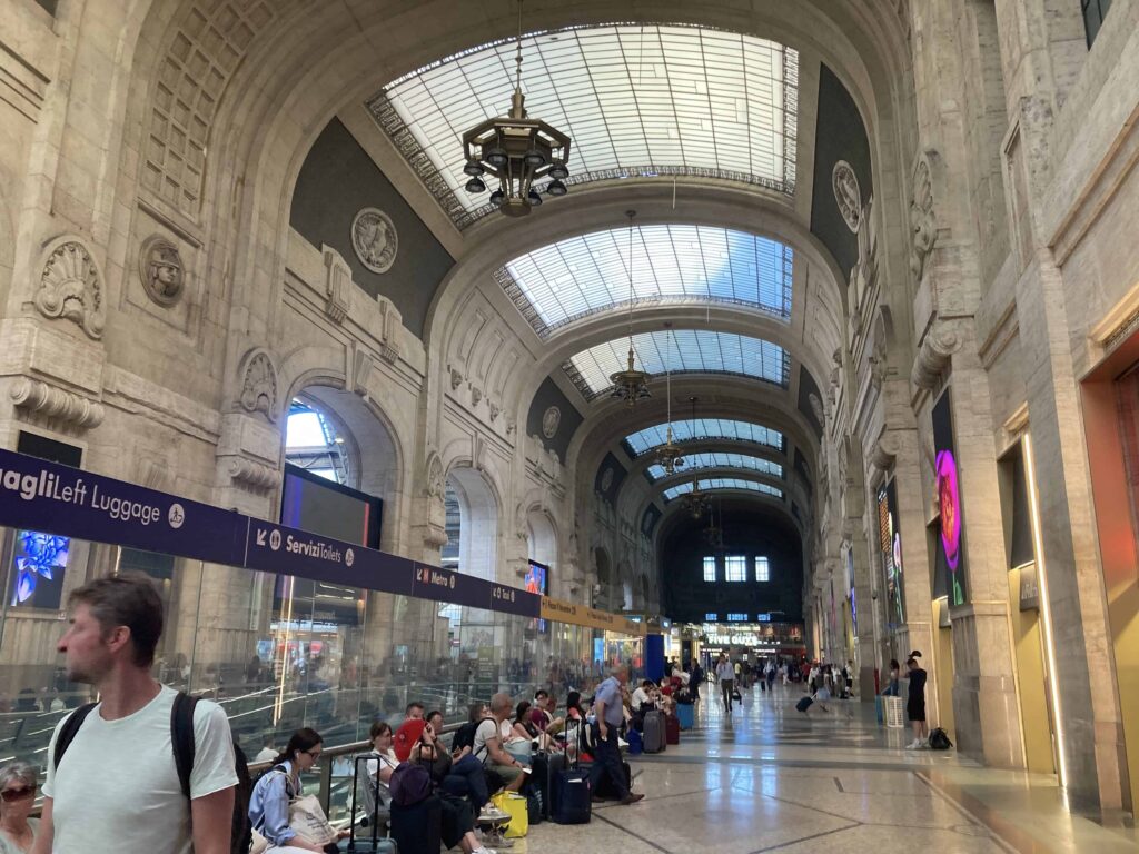 People sitting and waiting inside grand hall of Milano Centrale train station. Huge skylights in ceiling, stone building.