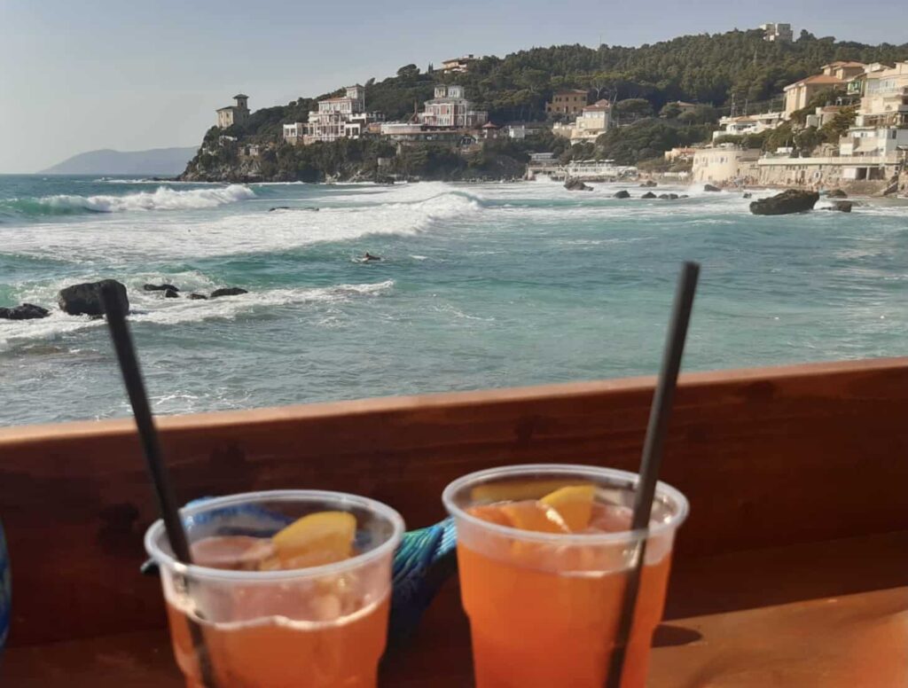 Two cups of Aperol spritz in the foreground with the waves crashing along the beach in Castiglioncello, Tuscany in the background. You can also see homes and wooded land in the background.