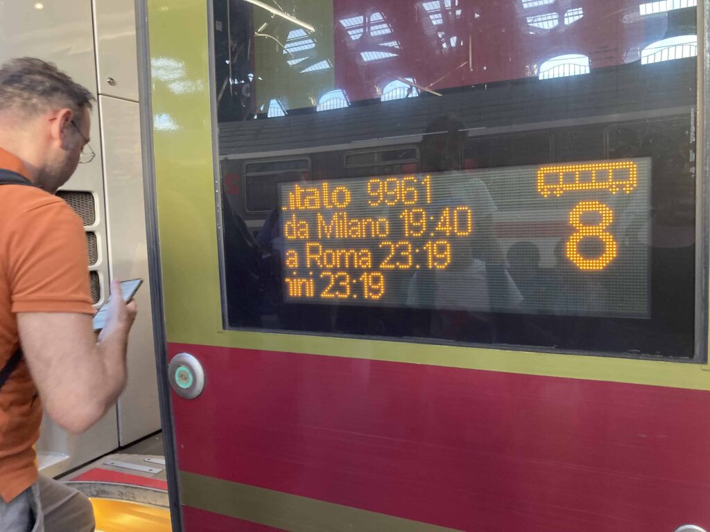 Man checking phone in front of train door in Italy. Digital sign with train info.