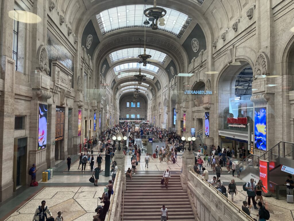 View of the large main hallway in the Milan train station. You can see a wide set of stairs, crowds of people walking around, digital train boards, and shops.
