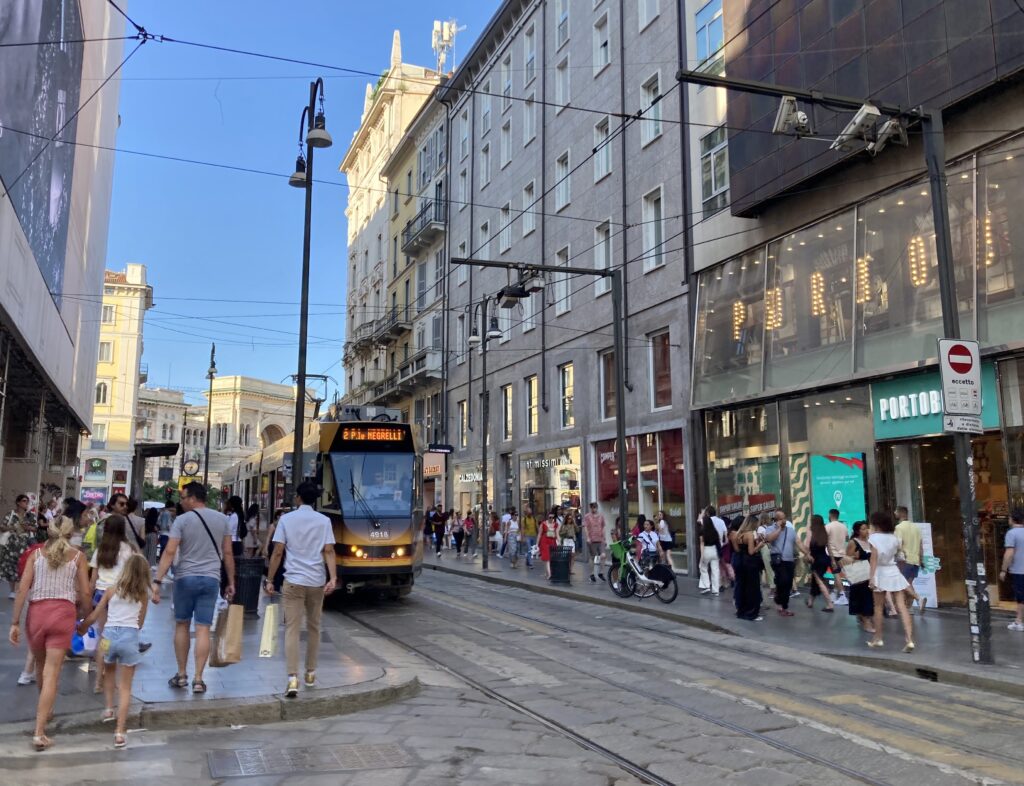 Tram moving down a street in Milan. People walk on sidewalks on both sides.