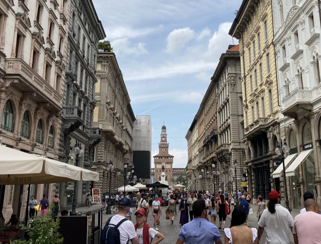 People walking on a pedestrian street in Milan, Italy. You can see Castello Sforzesco in the background.