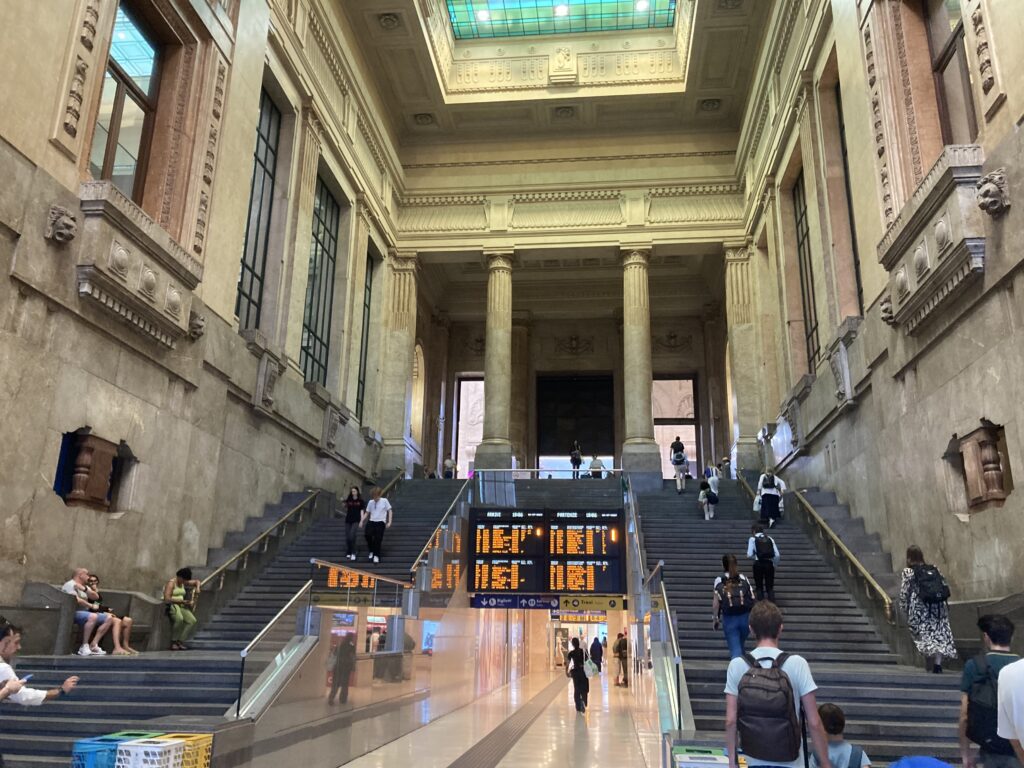 Two sets of stairs lead up to the first floor of the Milan train station. Another hallway also leads straight ahead toward digital train departure boards.