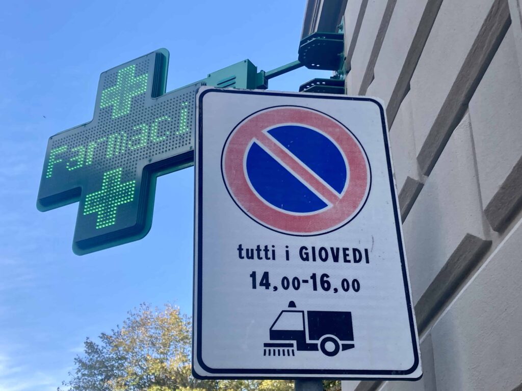 Green cross pharmacy sign in Italy behind a no parking and street sweeping sign.