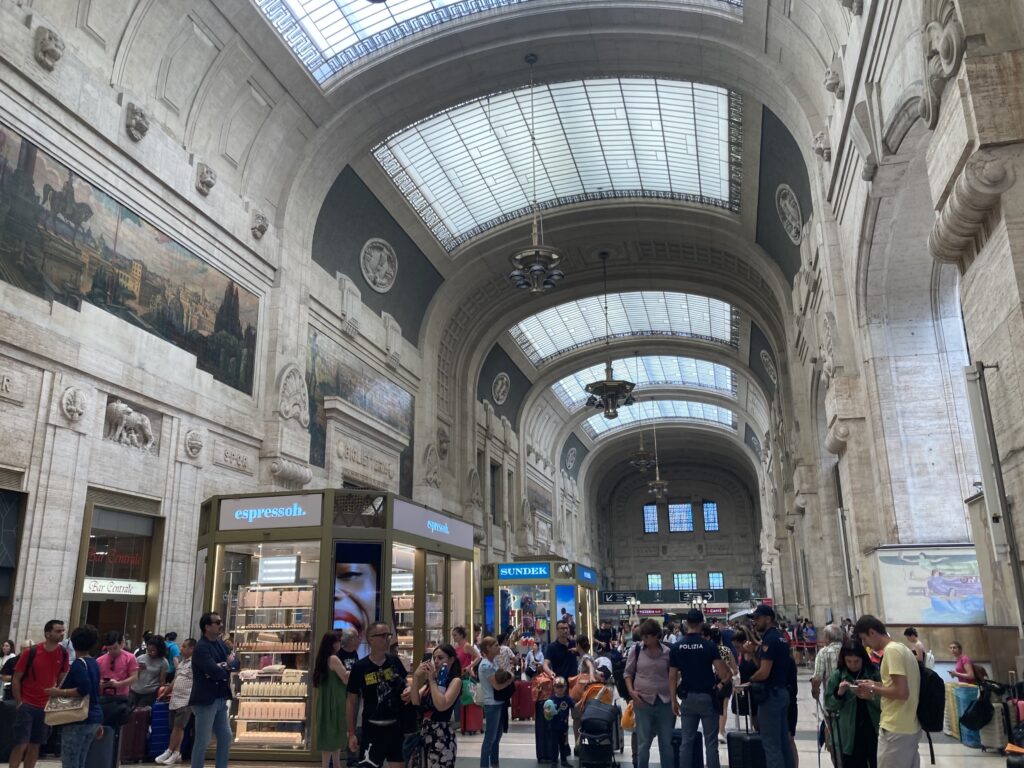 Light comes into the main hallway of the stone train station in Milan. People are in the hallway and you can see kiosks.