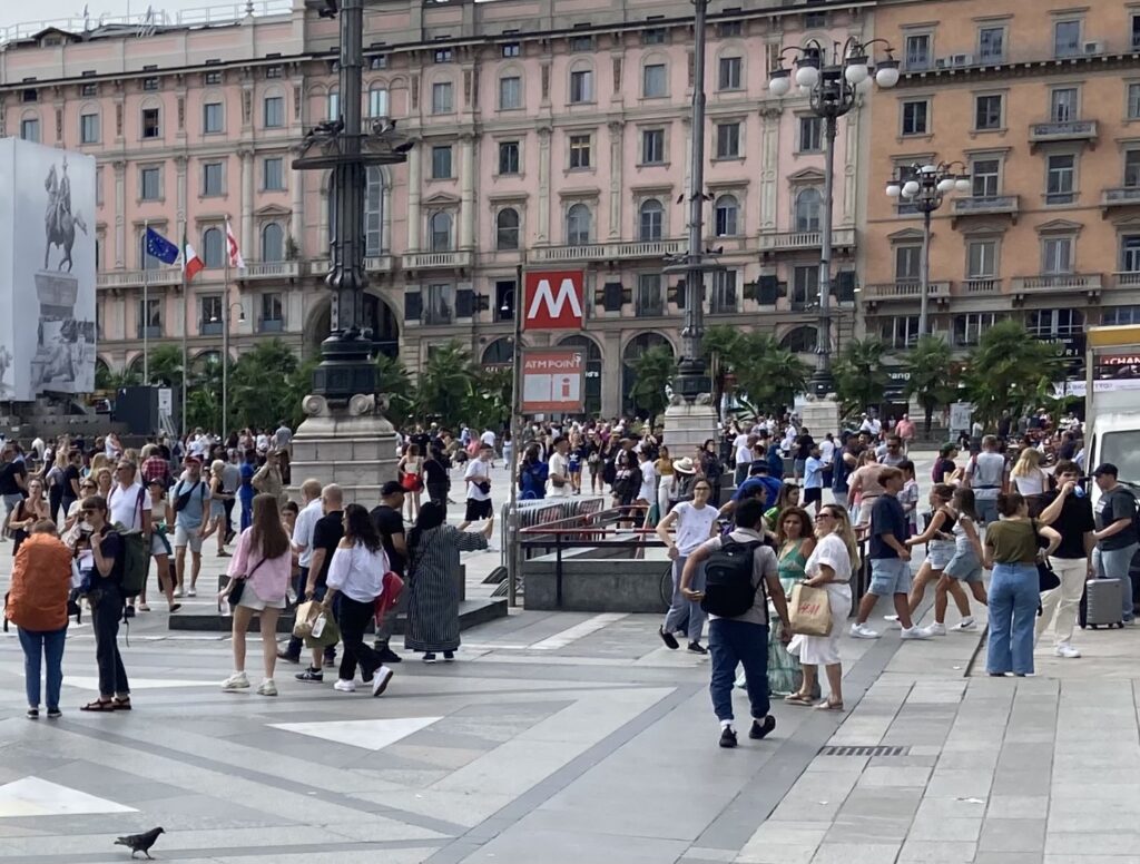 Red sign with white 'M' designates a metro underground station in Milan, Italy. People wait around outside in the Piazza Duomo.