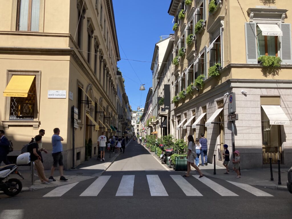 People crossing the street in Milan. Street sign on building on left for Via Monte Napoleone.