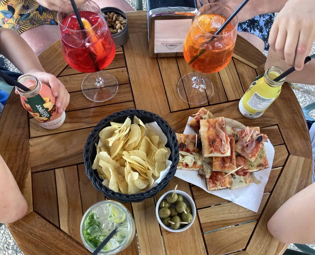 View of wooden table with people sipping drinks and eating snacks for aperitivo. Bowls of chips and olives and squares of pizza.